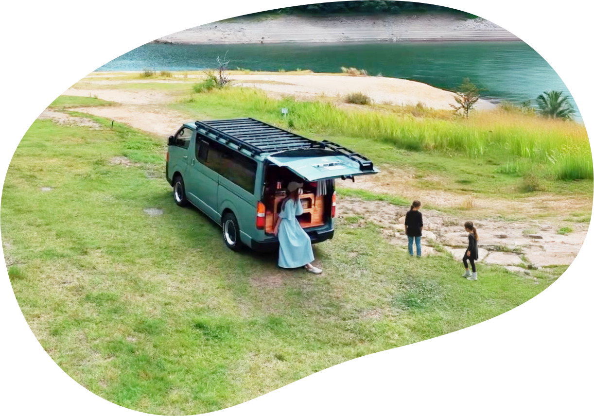 湖畔の草地にリアゲートを開けて停まるハイエース。女性と子どもたちが水辺で遊んでいる。